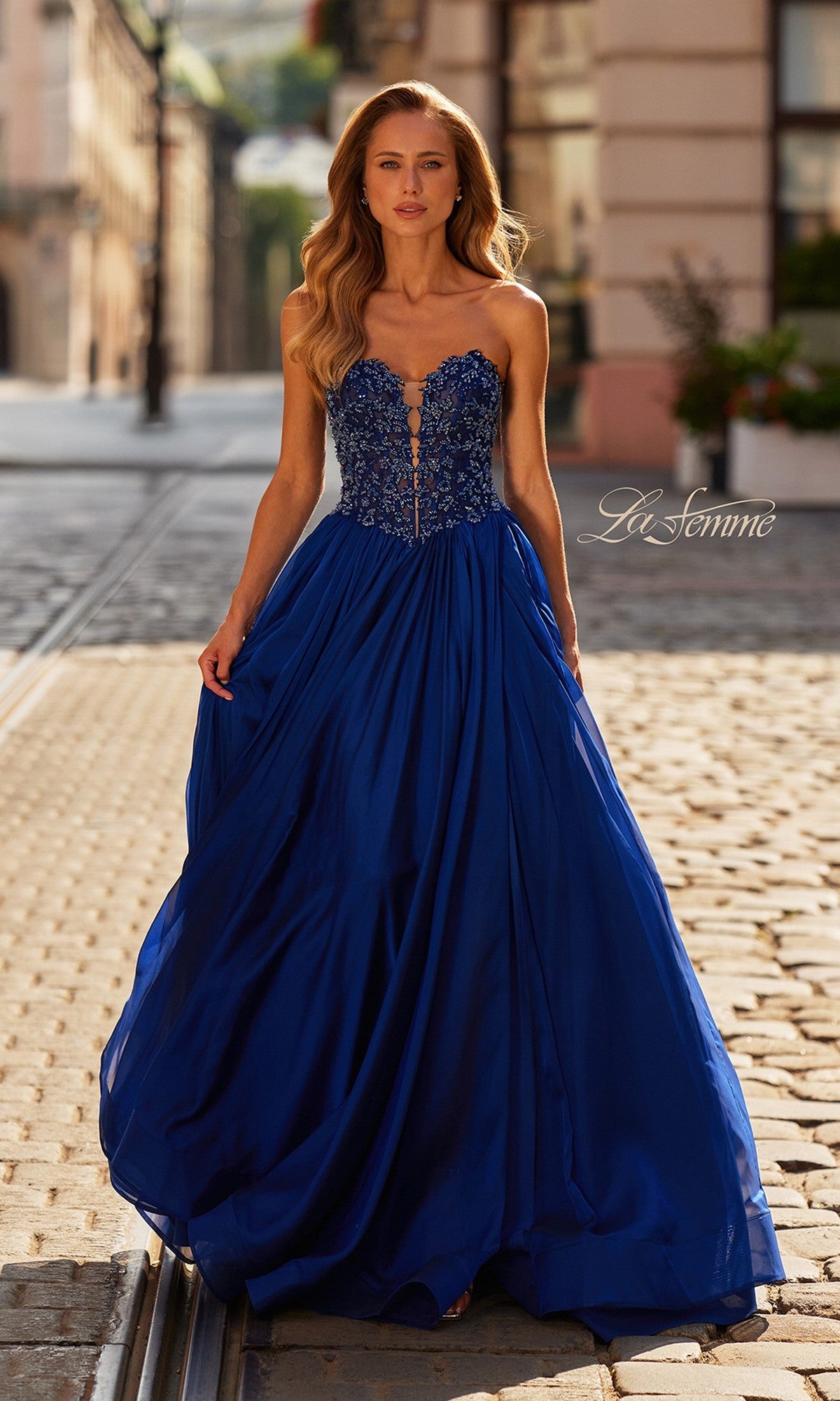 Wearing the La Femme 33552 long prom dress, a woman in a strapless chiffon gown with a beaded bodice and high leg slit stands gracefully on a cobblestone street in daylight.